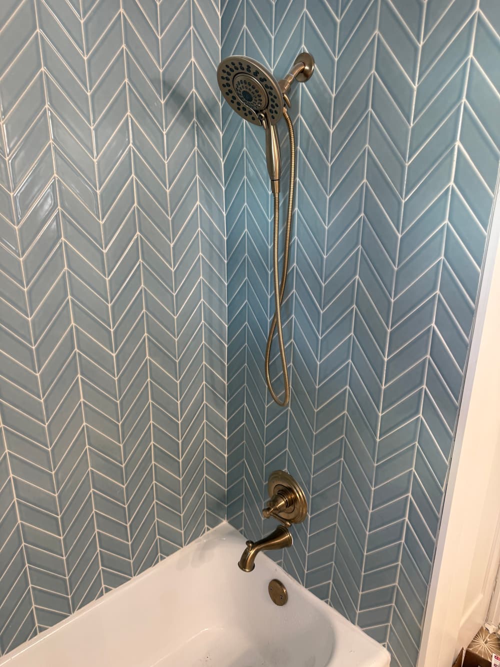 Modern bathroom shower fixture against blue herringbone tile wall with white bathtub.